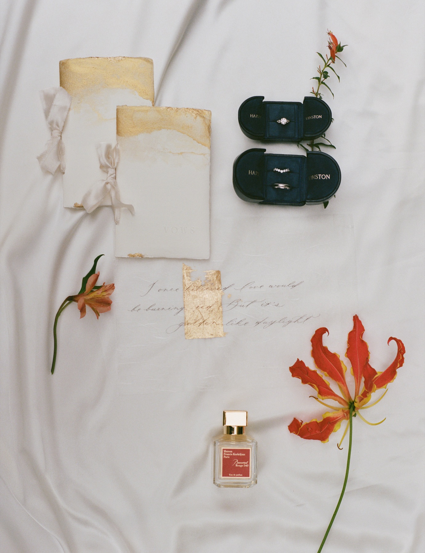 Wedding vows, rings, perfume, and red flowers arranged on soft white fabric.